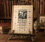 A vintage book titled 'Intérieurs Rustiques' by Charles-Moreau and Charles-Brun, displayed upright on an ornate metal book stand on a wooden table, with antique books and a ceramic vase in the background.