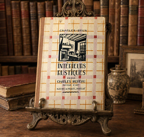 A vintage book titled 'Intérieurs Rustiques' by Charles-Moreau and Charles-Brun, displayed upright on an ornate metal book stand on a wooden table, with antique books and a ceramic vase in the background.