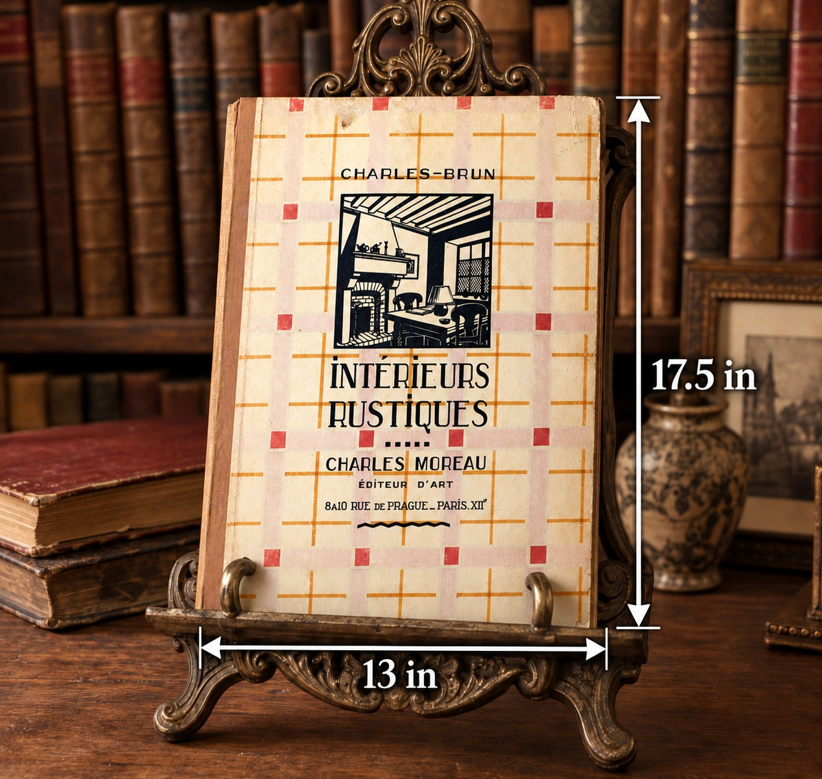 A vintage-style book display stand holding a book titled 'Intérieurs Rustiques' by Charles-Moreau, with overlaid dimension arrows showing 13 inches wide and 17.5 inches tall, set against a background of bookshelves.