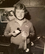 Gregg Ewell holding a duck carving and a large ribbon in an indoor setting with a poster of a duck on the wall.