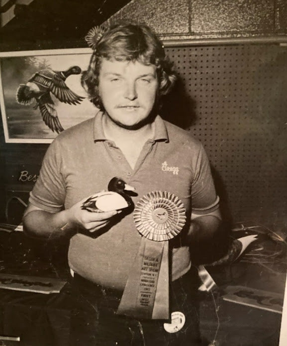 Gregg Ewell holding a duck carving and a large ribbon in an indoor setting with a poster of a duck on the wall.