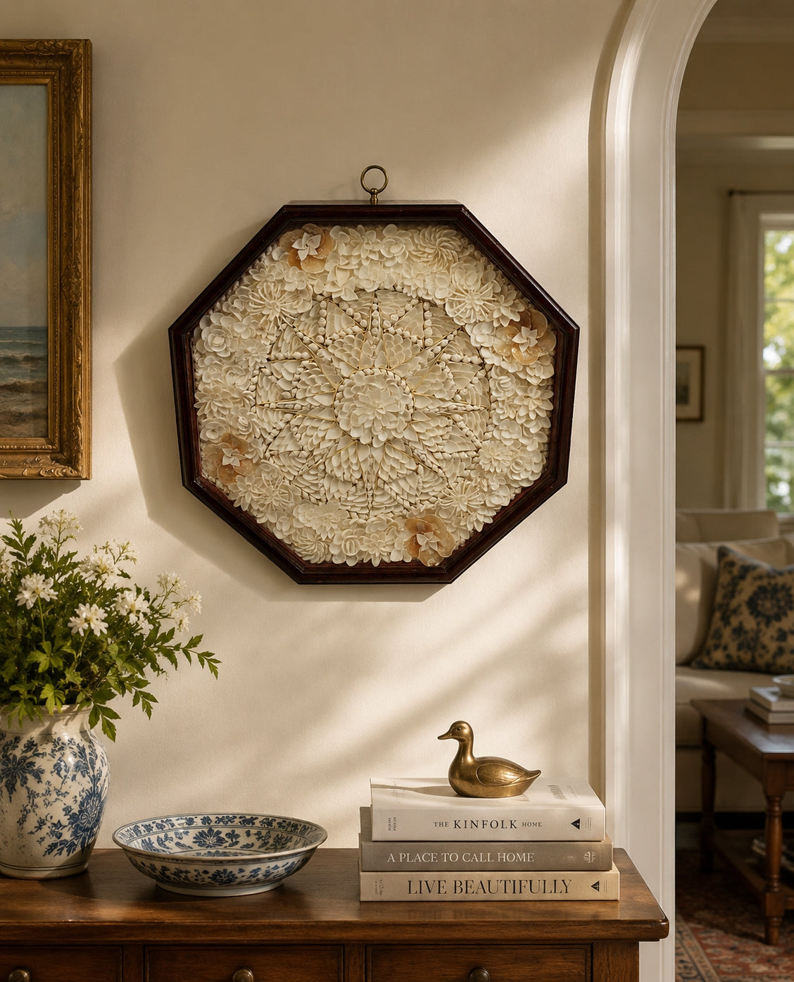 Decorative Sailors Valentine wall piece on a wall with a wooden side table below containing books, a vase, and a small sculpture.