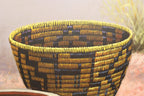 A close-up of a woven basket with a geometric pattern in blue, yellow, and brown, shown against a soft, painterly background.