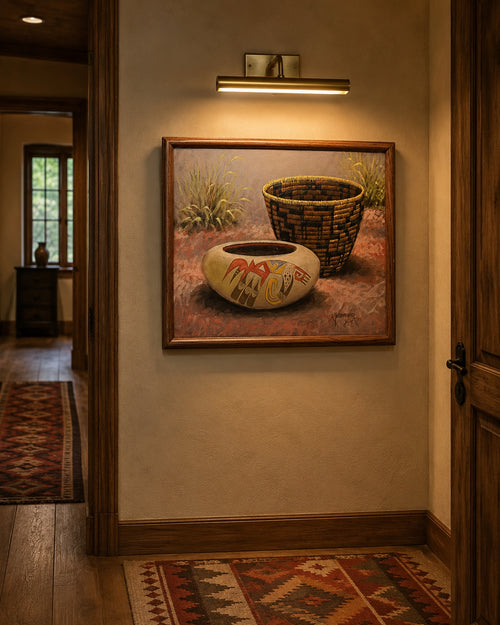 A framed art print depicting a Native American style pot and basket, displayed on a warm beige wall in a hallway with wood trim and a wall sconce above. By Yellowhair