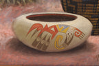 A painted ceramic pot with a cream-colored body and colorful geometric and animal-inspired motifs in red, yellow, brown, and blue, set against a textured reddish-brown background.