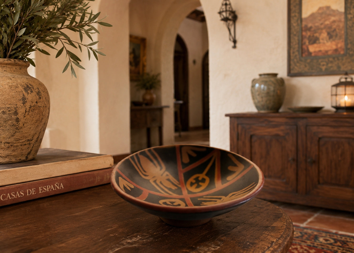 A hand-painted ceramic bowl with a black base and warm orange geometric patterns sits on a rustic wooden table in a warm, traditional Spanish-style room. Peruvian Art Pottery Terra Cotta Bowl with Nazca Spider Design