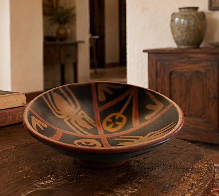 A hand-painted ceramic bowl with geometric and spider motifs in terracotta and ochre on a dark background, displayed on a rustic wooden table in a warm, traditional room.