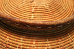 Close-up of a hand-woven basket with concentric circular patterns in natural tan, brown, and orange tones, showing the detailed texture of the woven material and a dark brown rim.