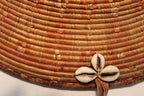 Close-up of a woven basket made from tightly coiled natural fibers in warm tan and brown tones, decorated with a small cluster of cowrie shells at the front.