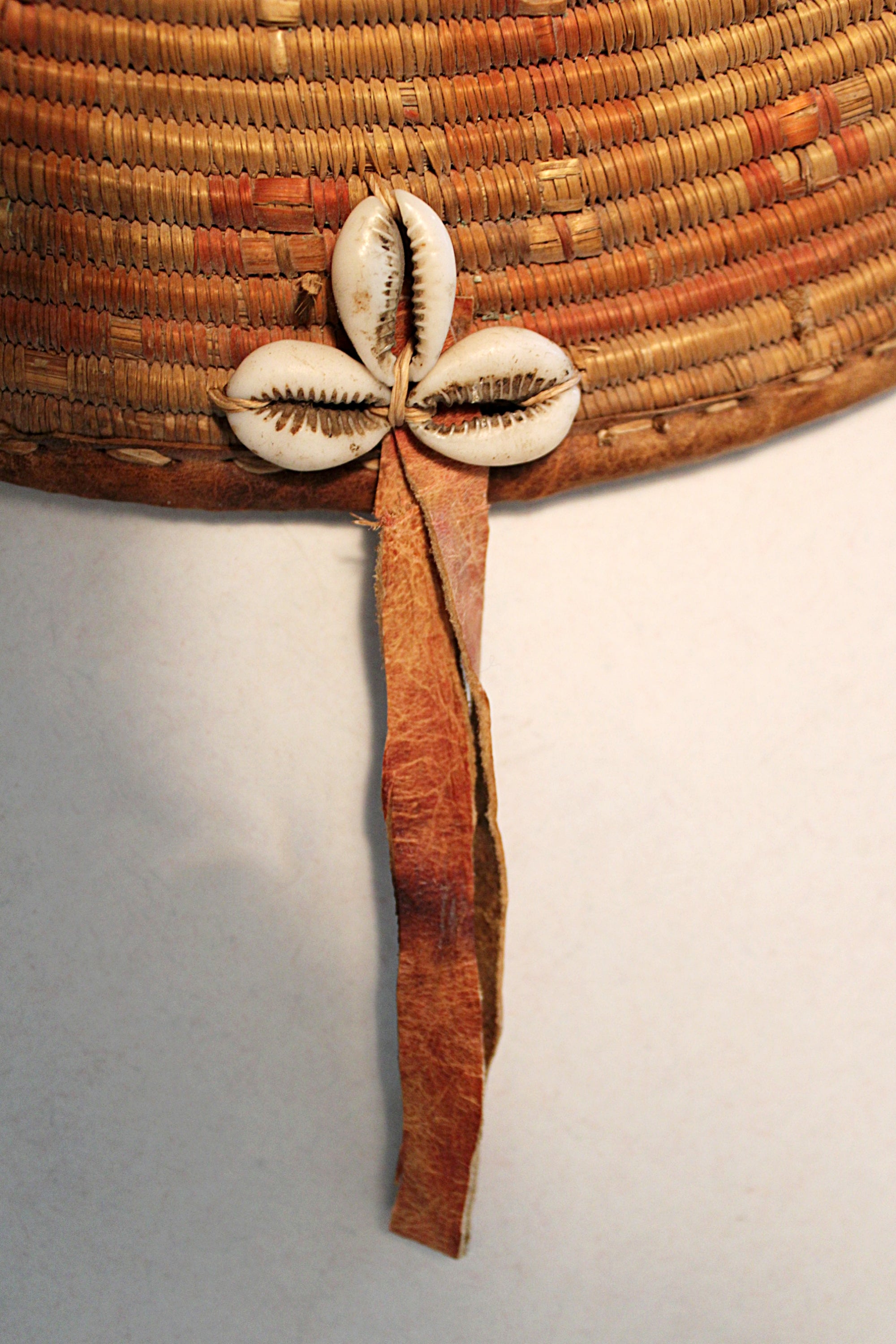 A close-up of a woven basket in natural tan and brown tones, featuring a decorative cluster of cowrie shells and a long brown leather ribbon hanging down against a plain light background.