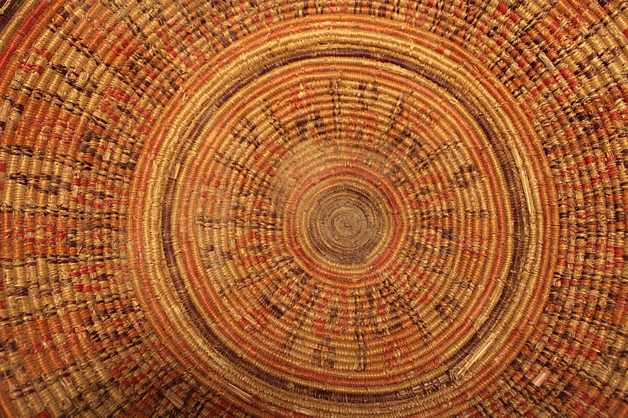 A close-up of a circular woven basket with concentric rings in natural tan, brown, orange, and red tones, filling the entire frame.