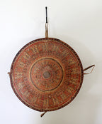 A round woven basket with concentric circular patterns in red, brown, and beige, hanging from a black hook against a plain white wall.