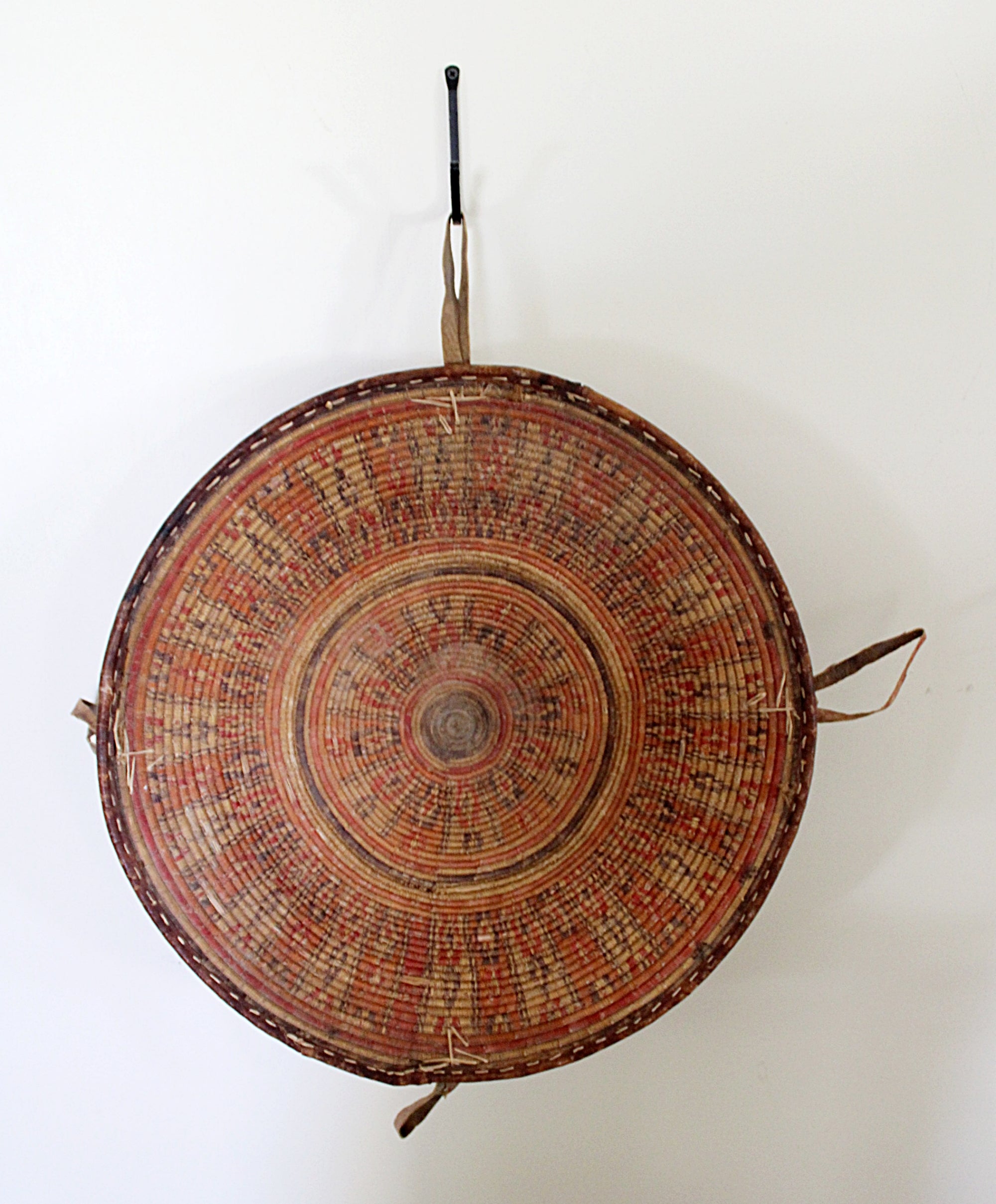 A round woven basket with concentric circular patterns in red, brown, and beige, hanging from a black hook against a plain white wall.
