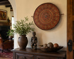A round woven basket on wall with intricate red, brown, and beige concentric patterns is displayed on a cream-colored wall above a dark wood console table. The table is styled with a large brown ceramic vase containing green foliage, a small dark wooden figurine, and a matching dark wood bowl with decorative woven spheres.