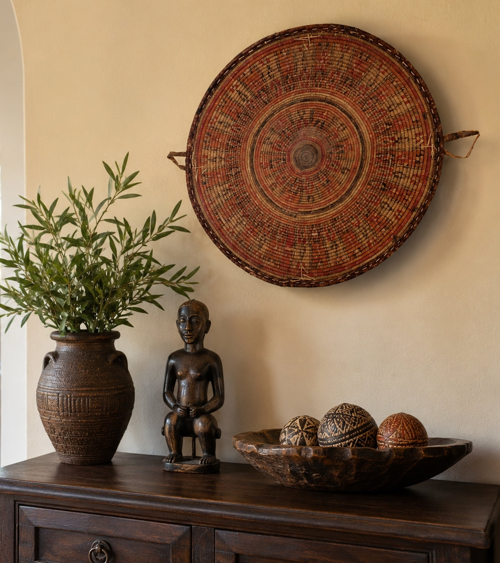 A large circular woven wall basket hanging with geometric patterns in earthy red, brown, and beige tones, displayed on a beige wall above a dark wooden console table decorated with a potted plant, a small statue, and a bowl of woven ornaments.