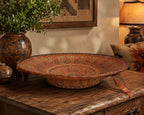 A round woven basket tray with a raised base, decorated with intricate red, orange, and brown geometric patterns, displayed on a rustic wooden table in a warm, styled room.