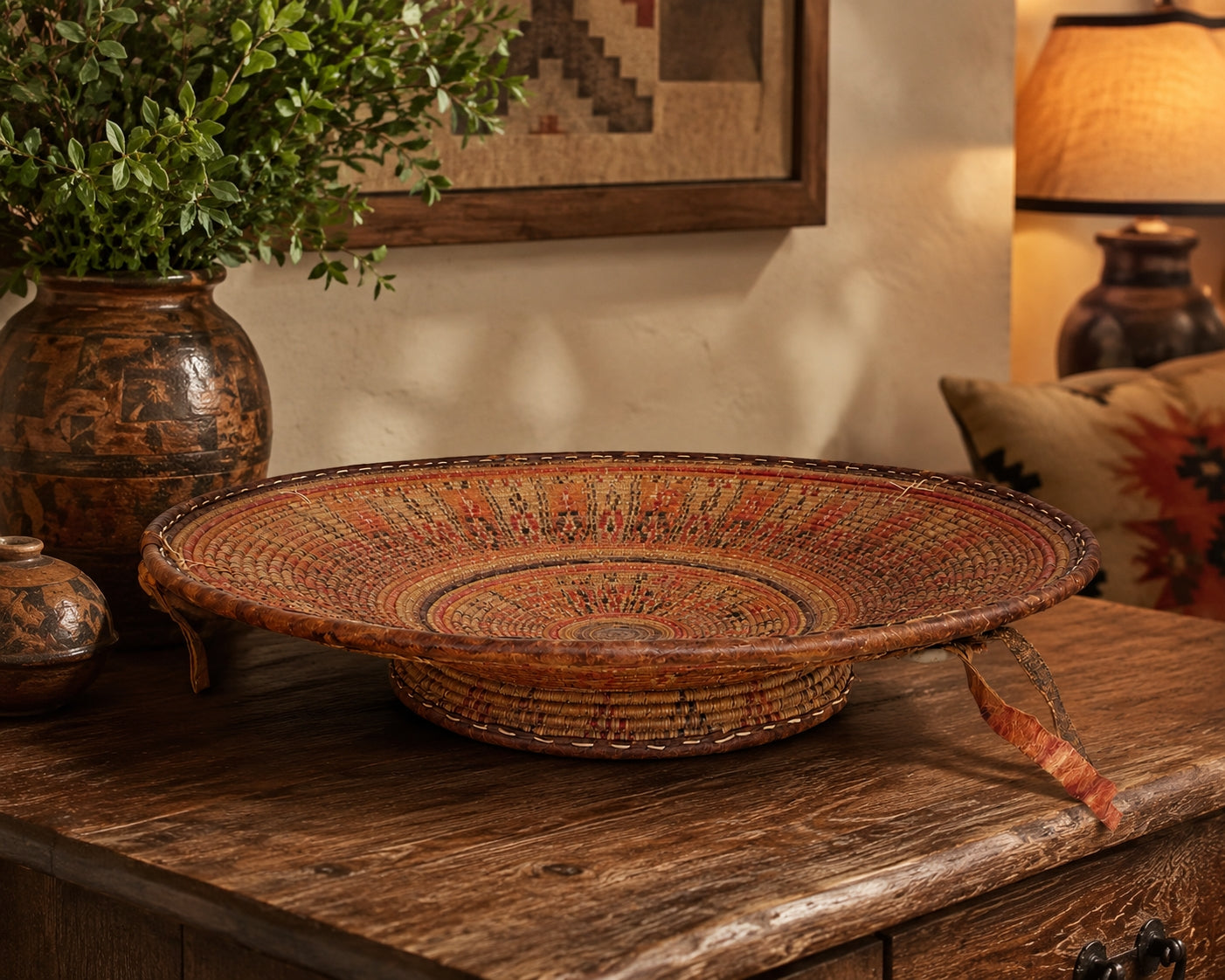 A round woven basket tray with a raised base, decorated with intricate red, orange, and brown geometric patterns, displayed on a rustic wooden table in a warm, styled room.