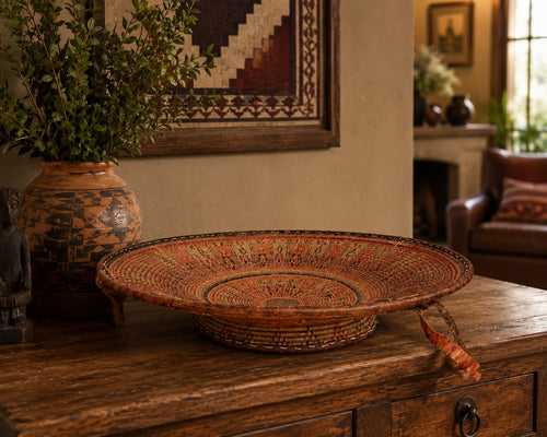 A large, round hand-woven basket bowl with intricate geometric patterns in earthy red, brown, and beige tones, displayed on a rustic wooden console table in a warm, traditional room setting.