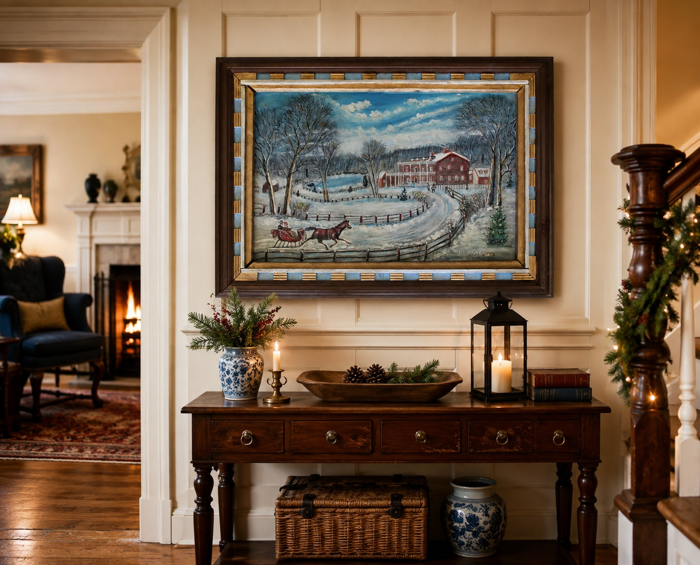 A framed winter landscape painting hangs on a cream paneled wall above a dark wood console table decorated with candles, a basket, and ceramic vases in a cozy home interior.
