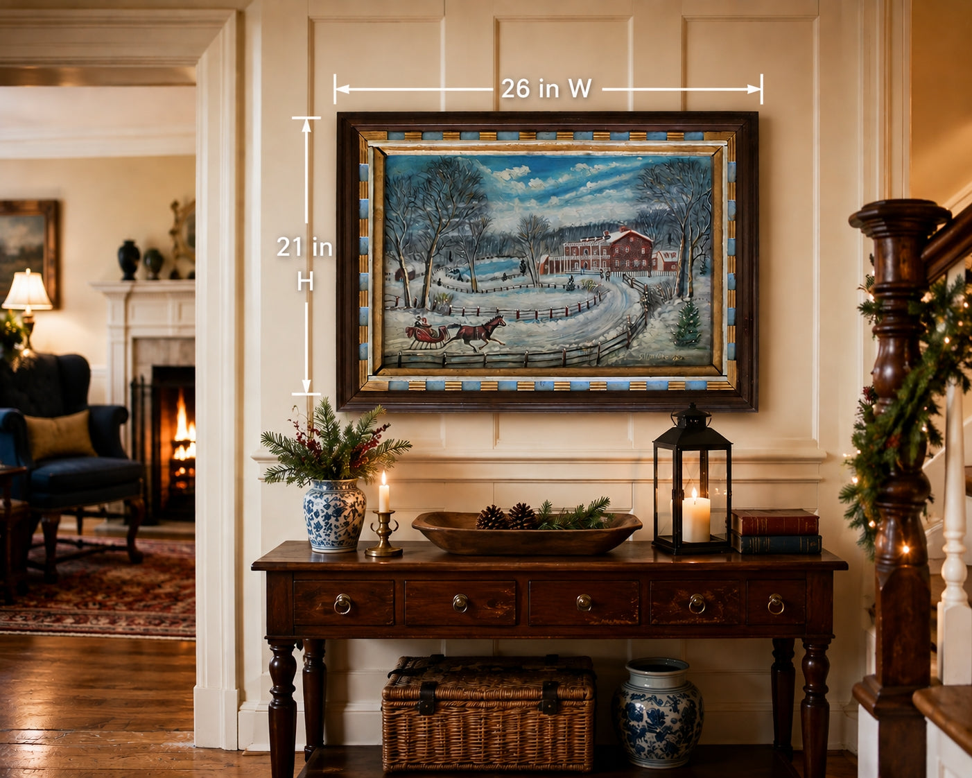 A framed winter landscape painting hangs on a beige paneled wall above a wooden console table in a decorated home interior. White dimension lines indicate the artwork is 26 inches wide by 21 inches high.