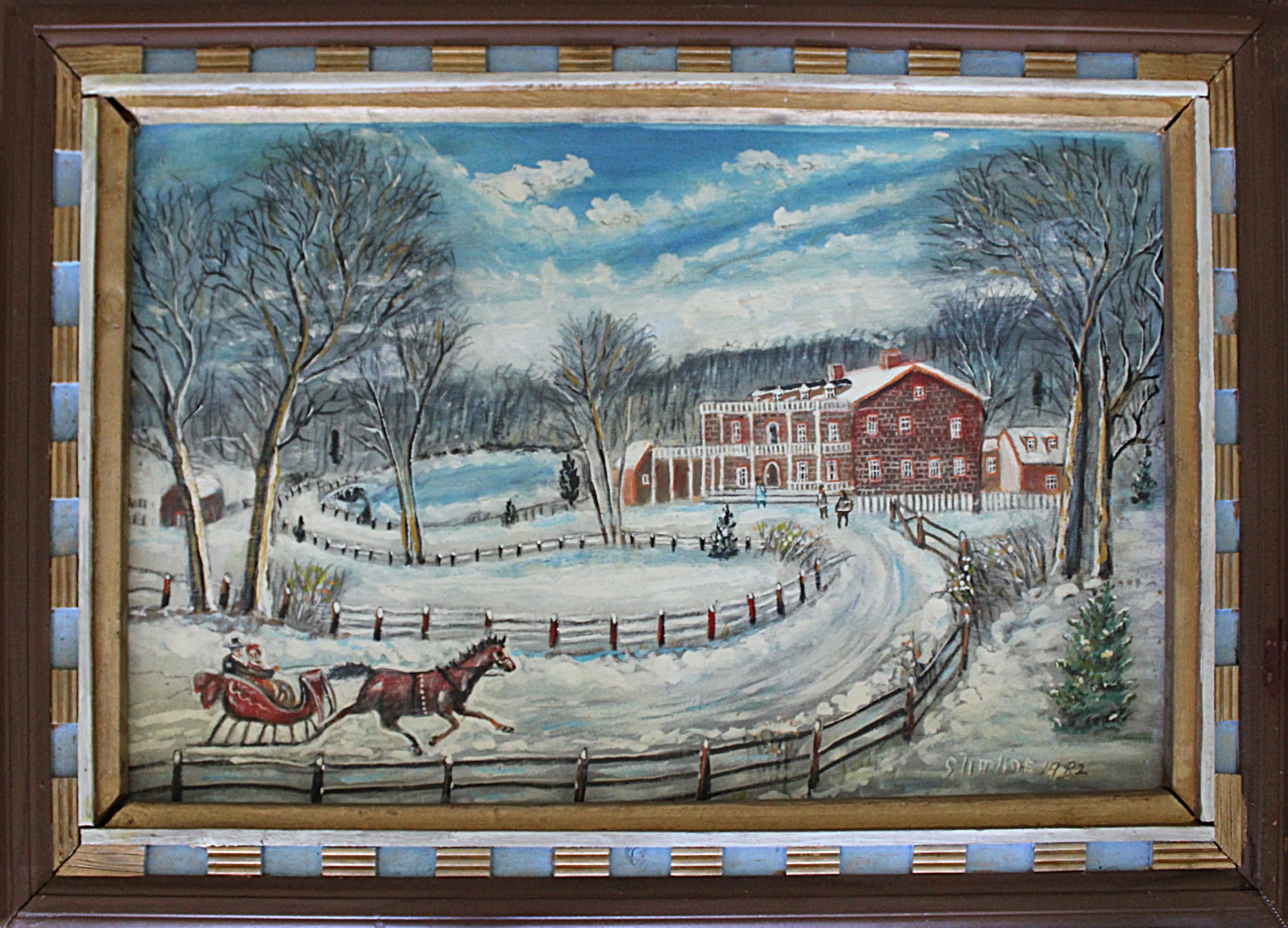 A framed winter landscape painting depicting a snowy farm scene with bare trees, a red barn, a sleigh with a horse, and a blue sky with clouds. The painting is housed in a dark brown wooden frame with a blue and gold striped inner border.