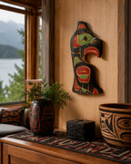 A hand-carved wooden wall art piece with Indigenous-style totem designs in red, green, and black, displayed on a wooden wall in a rustic room setting with a window showing a lake view.